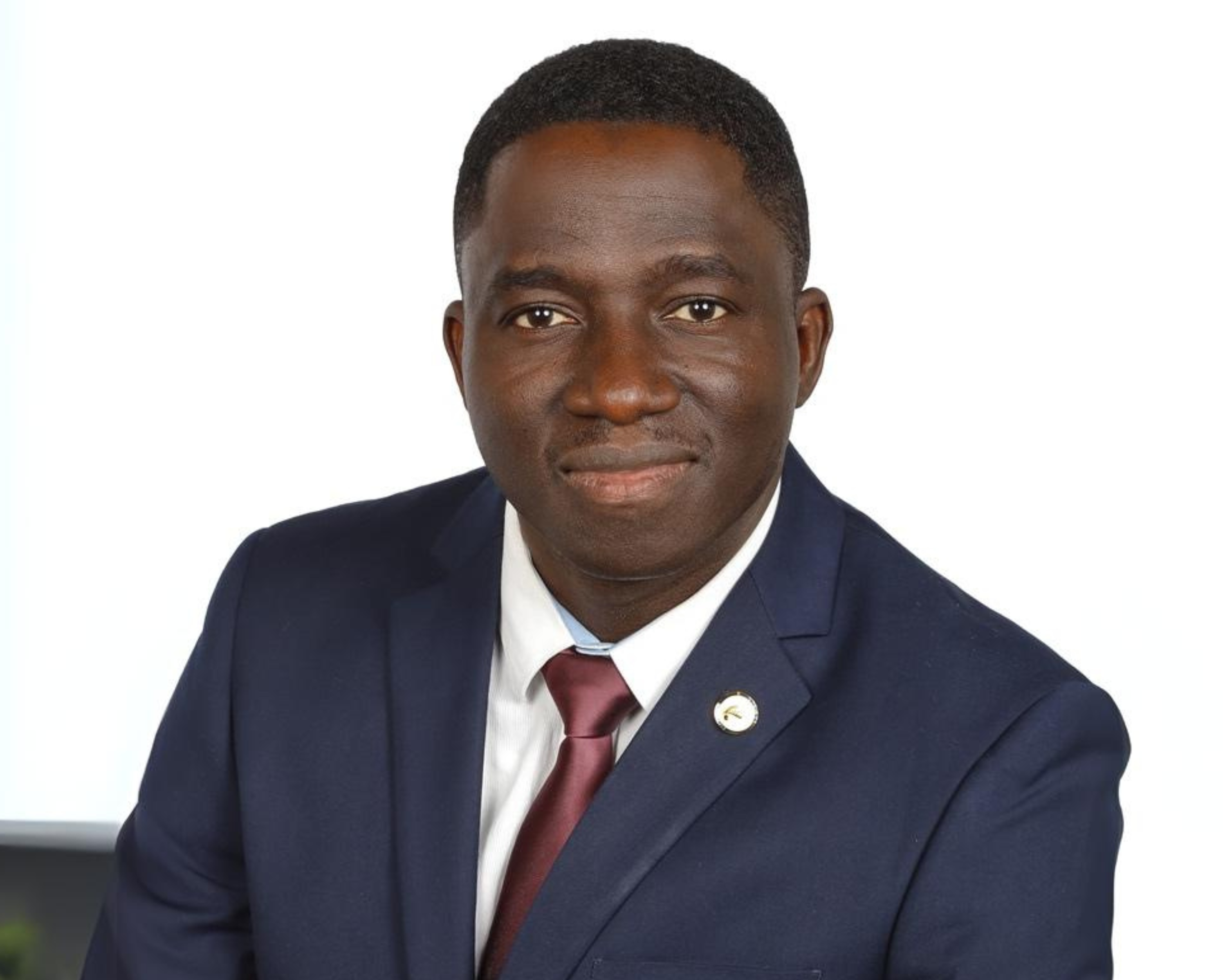 A portrait of a man wearing a dark blue suit, white shirt, and maroon tie, sitting against a white background. He has short, neatly trimmed hair and a calm, confident expression on his face. A small pin with a logo is visible on his left lapel.
