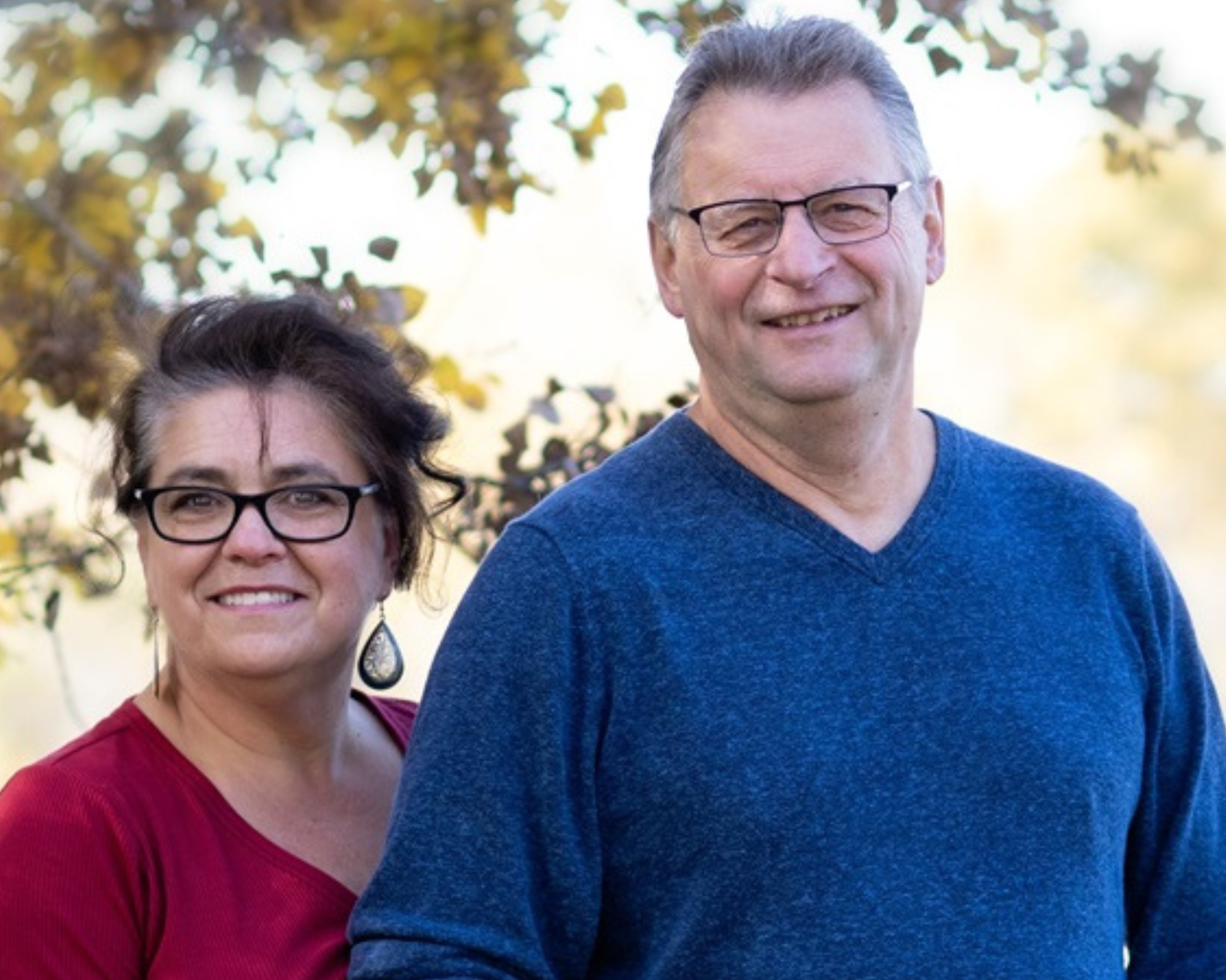 A couple standing outdoors with autumn leaves in the background. The woman is wearing a red top and glasses, while the man is wearing a blue sweater and glasses, both smiling warmly.