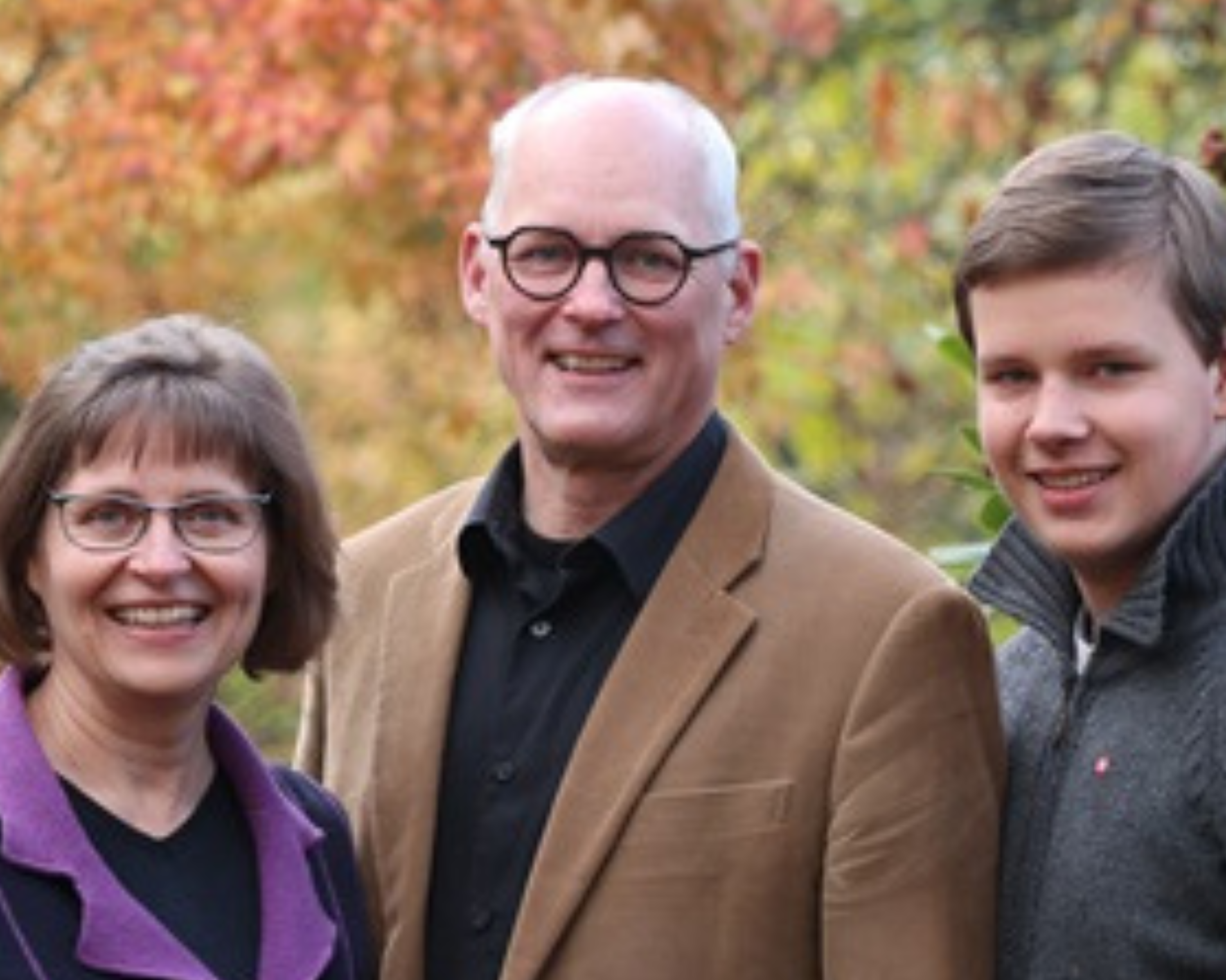 A family portrait outdoors with a middle-aged couple and a young adult male. The woman, wearing glasses and a purple cardigan, stands to the left. The man in the middle wears glasses, a tan jacket over a black shirt, and the young man on the right sports a grey sweater. They are all smiling, with autumn-coloured leaves in the soft-focus background.