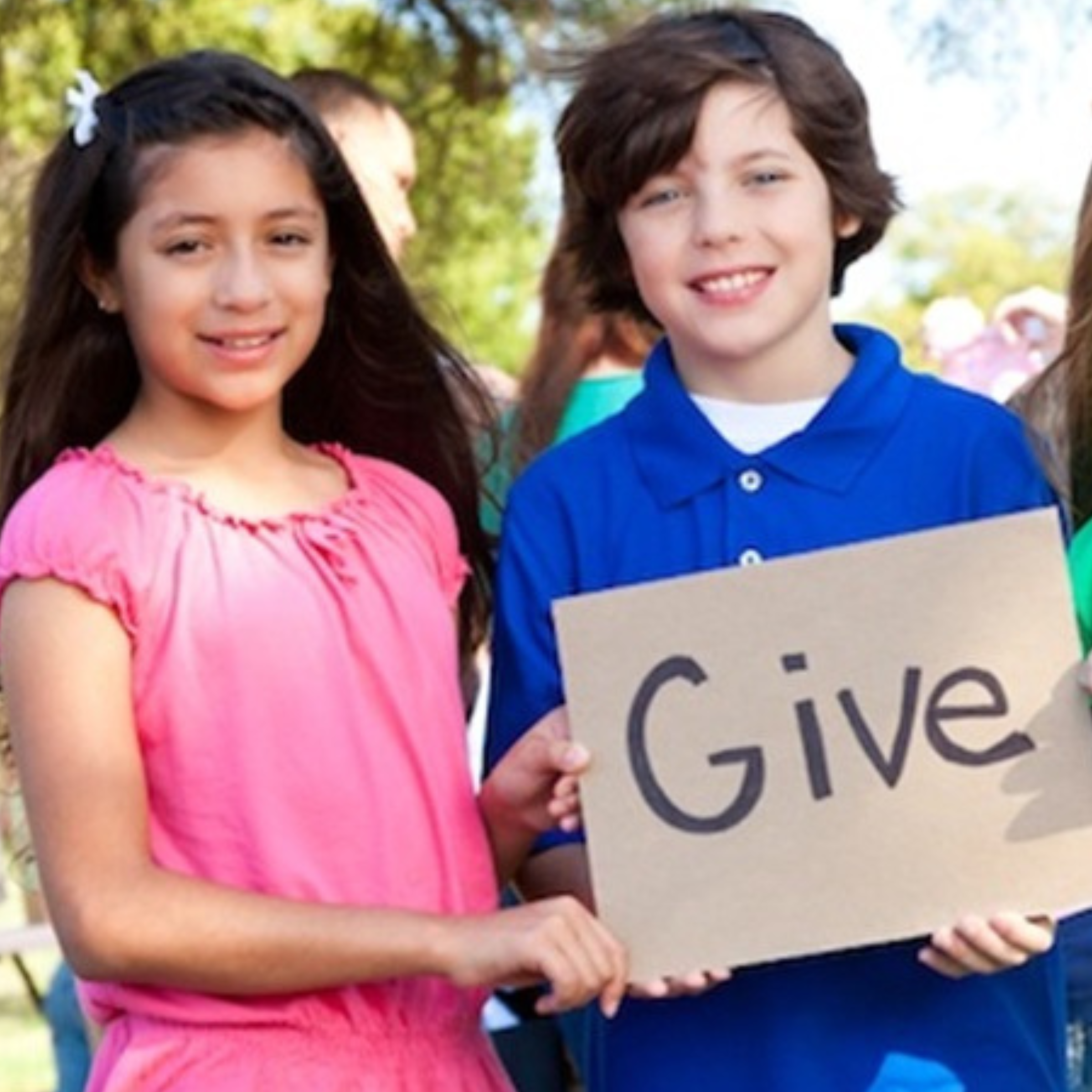 The image shows three smiling children standing outdoors, holding a cardboard sign that says "Give." The children are dressed in bright, colorful clothing—one girl in pink, a boy in blue, and another girl in green—emphasizing the cheerful and positive atmosphere. They are standing in a sunny park, with trees and other people visible in the background. The scene conveys a message of generosity, kindness, and community involvement, with the children promoting the idea of giving and helping others.