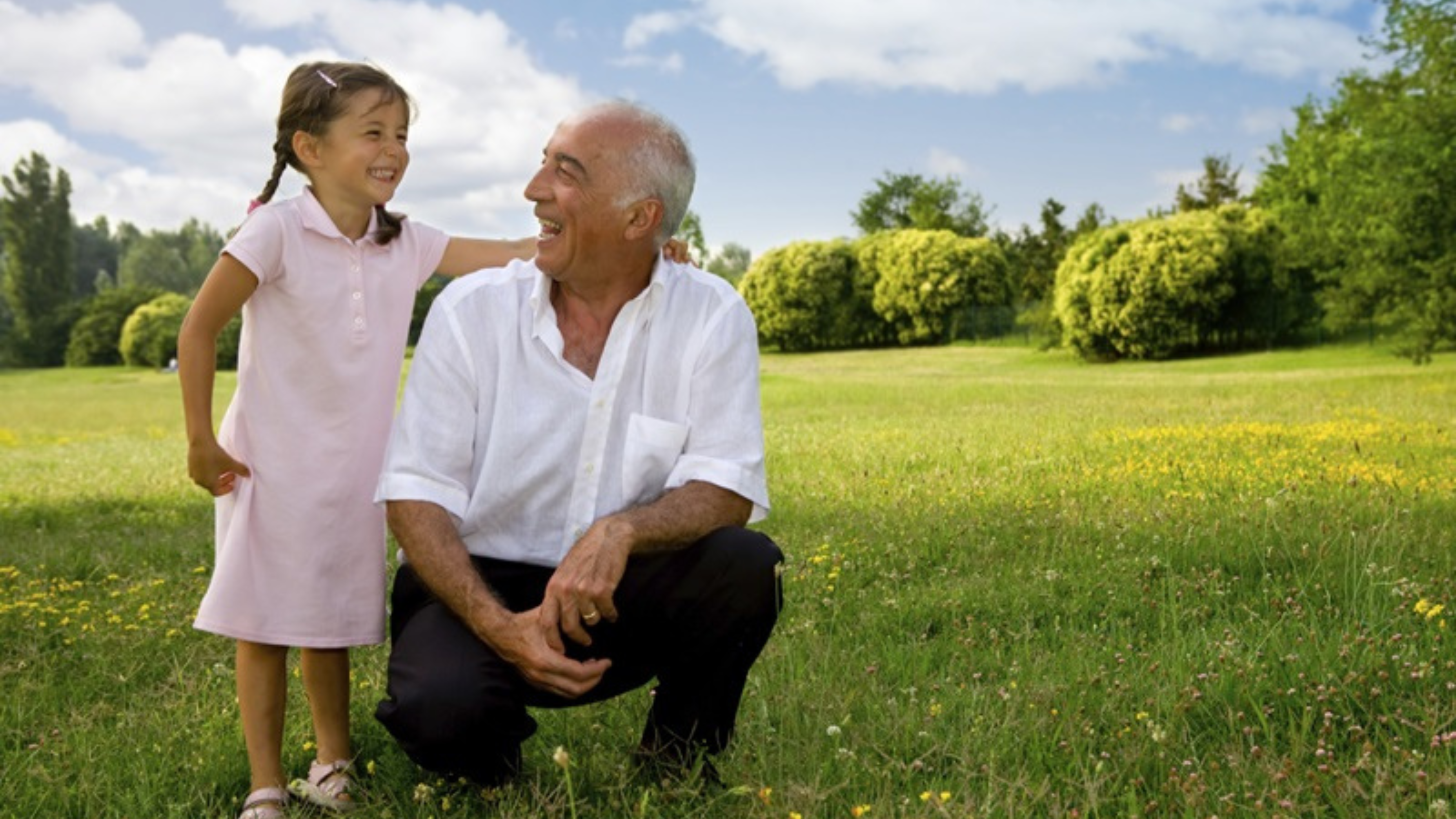 A heartwarming scene of a senior man and a young girl in a sunny park. The man, with grey hair and dressed in a white shirt, is kneeling on one knee, smiling and looking at the girl, who is standing next to him, wearing a pink dress and also smiling. They are surrounded by a lush green field dotted with yellow flowers, with trees and blue sky in the background.