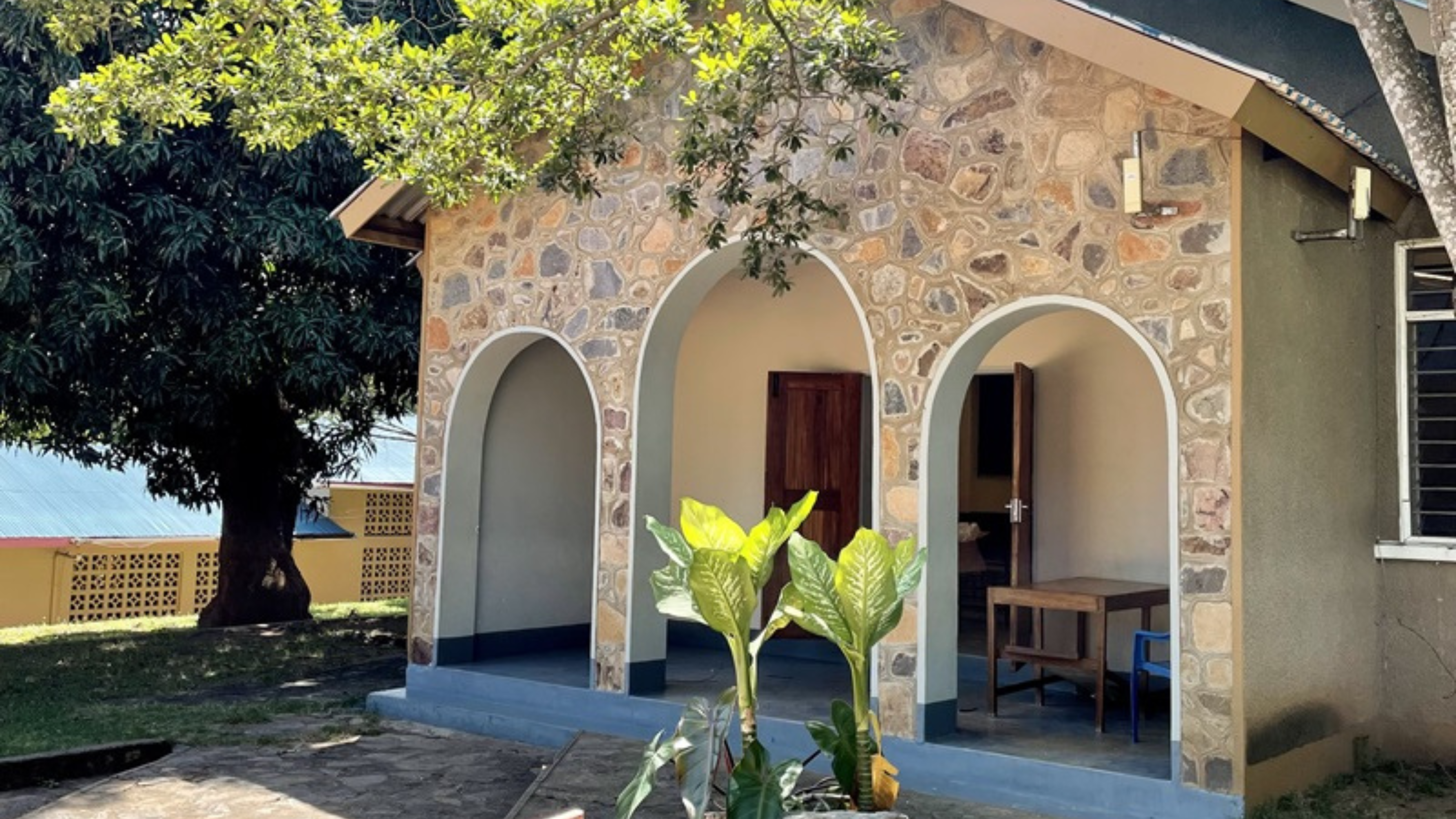 A quaint stone chapel under a clear blue sky, surrounded by lush green trees. A 'CHAPEL' sign points towards the open arched entrance, with a potted plant prominently displayed in front of the sign.
