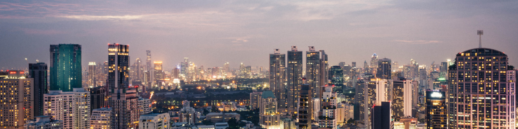 A panoramic view of a bustling city skyline at dusk, with towering skyscrapers illuminated by city lights under a purple-hued sky.