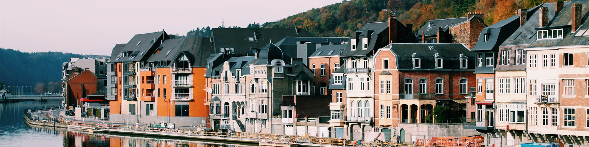 A row of colorful, traditional European-style buildings along a riverside. The buildings have varied architectural designs, with a mix of brick and stone facades, large windows, and balconies. The river runs alongside a walkway, and in the background, there are forested hills with autumn-coloured foliage. The scene is calm and picturesque, capturing a charming, small-town atmosphere. This photo is by Alex Vasey on Unsplash.