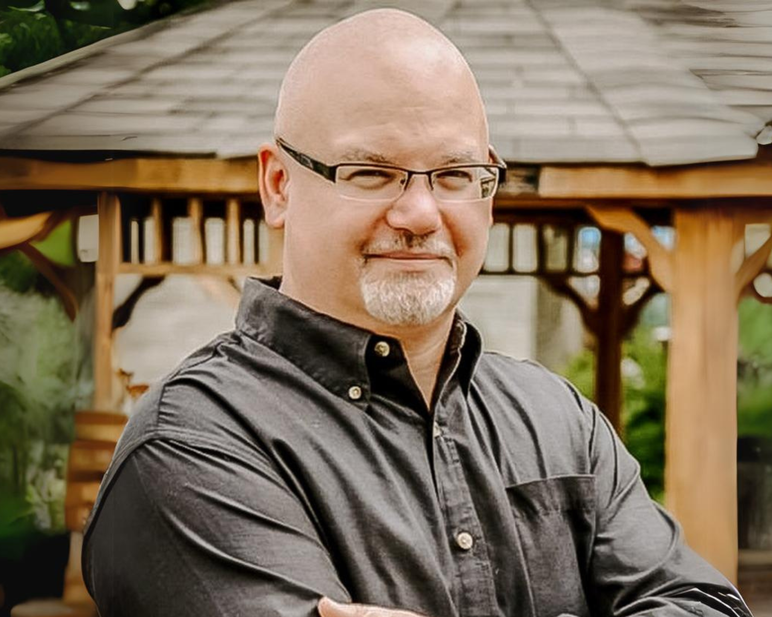 A man with a bald head and a short white beard is standing outdoors with his arms crossed, smiling confidently. He is wearing glasses and a dark button-up shirt. Behind him is a wooden gazebo, and the background features lush greenery, creating a peaceful and inviting atmosphere.