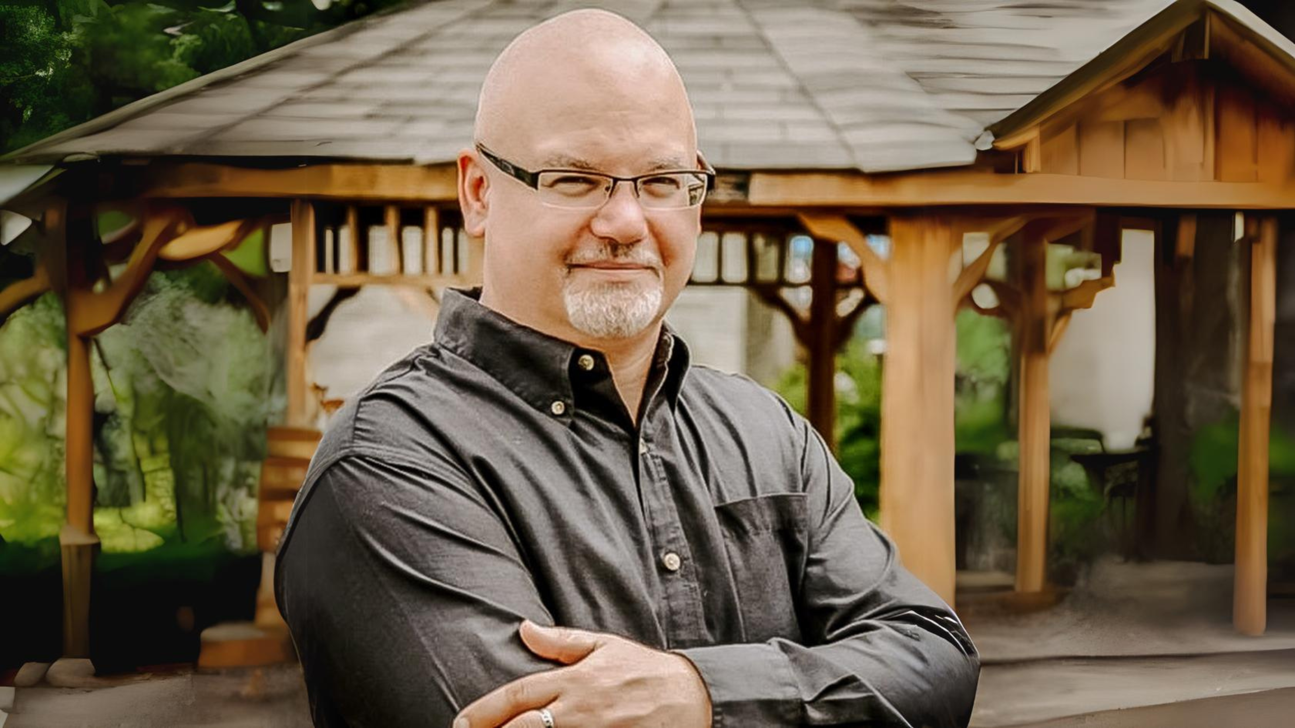 A man with a bald head and a short white beard is standing outdoors with his arms crossed, smiling confidently. He is wearing glasses and a dark button-up shirt. Behind him is a wooden gazebo, and the background features lush greenery, creating a peaceful and inviting atmosphere.