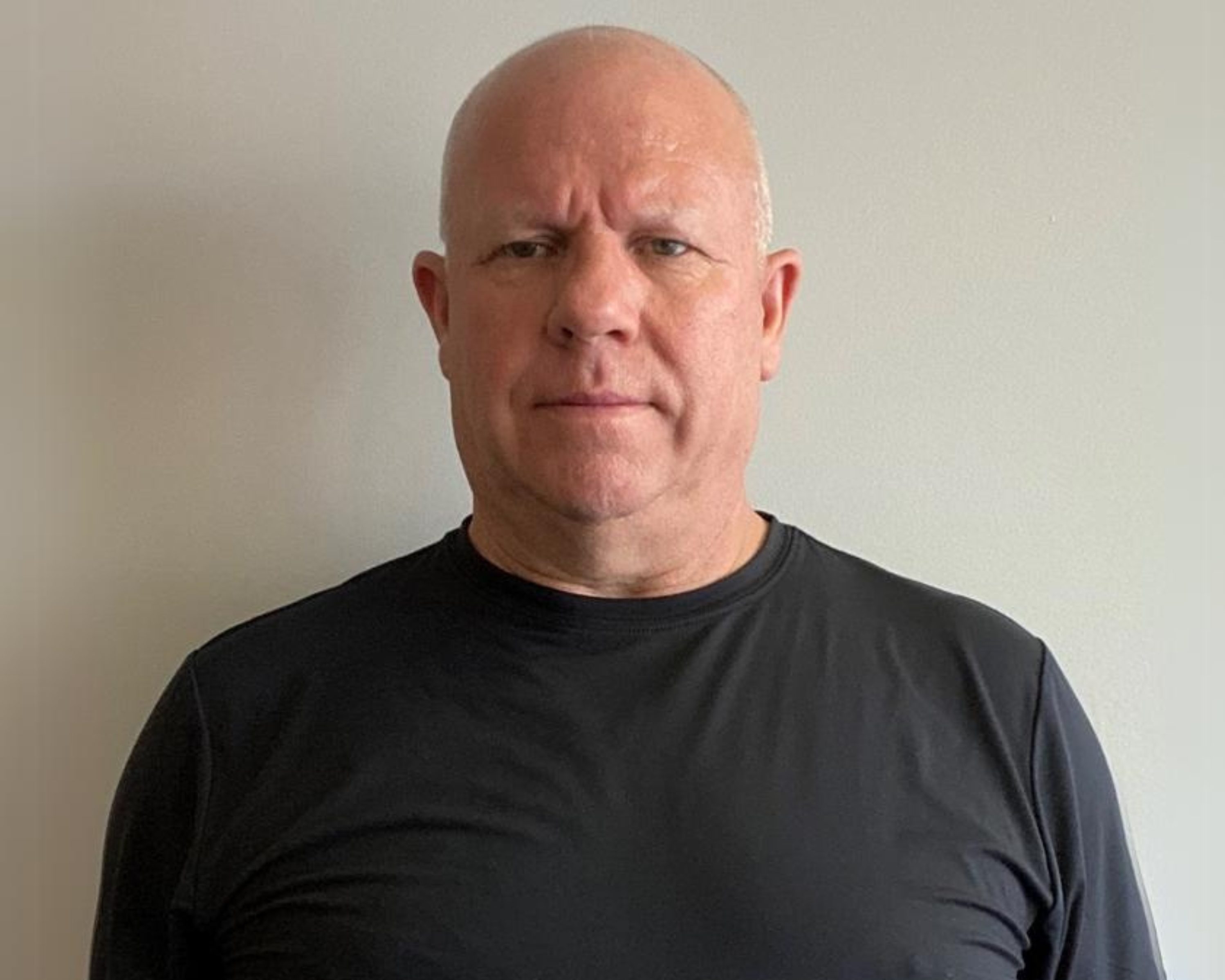 A serious-looking man with a bald head is standing against a plain, light-coloured wall. He is wearing a black t-shirt and looking directly at the camera. The photo is straightforward, focusing on his face and upper body.