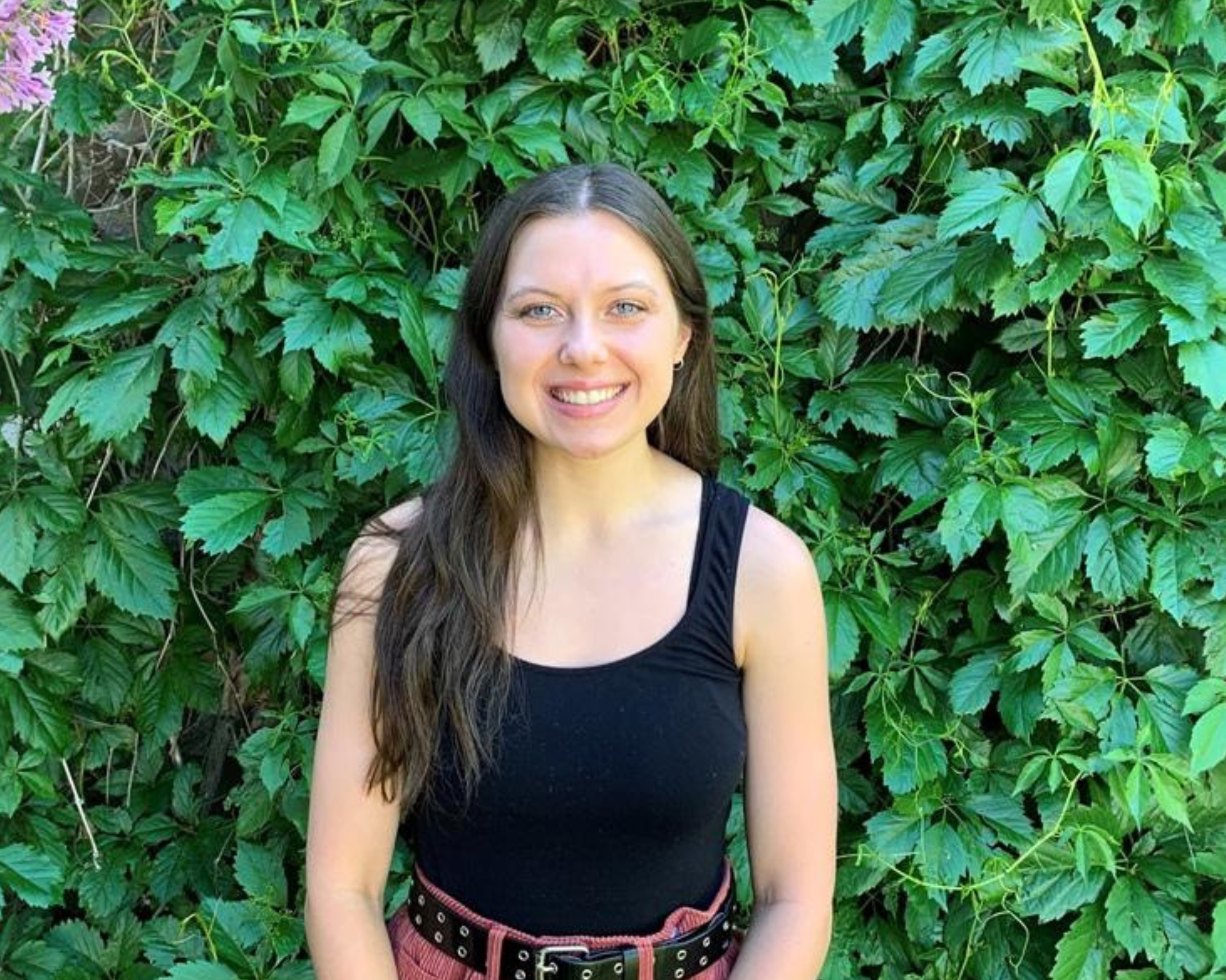 A young woman with long brown hair is smiling while standing in front of a lush green wall of leaves and plants. She is wearing a black sleeveless top and a belt with a red skirt. The vibrant greenery behind her creates a natural and serene backdrop.