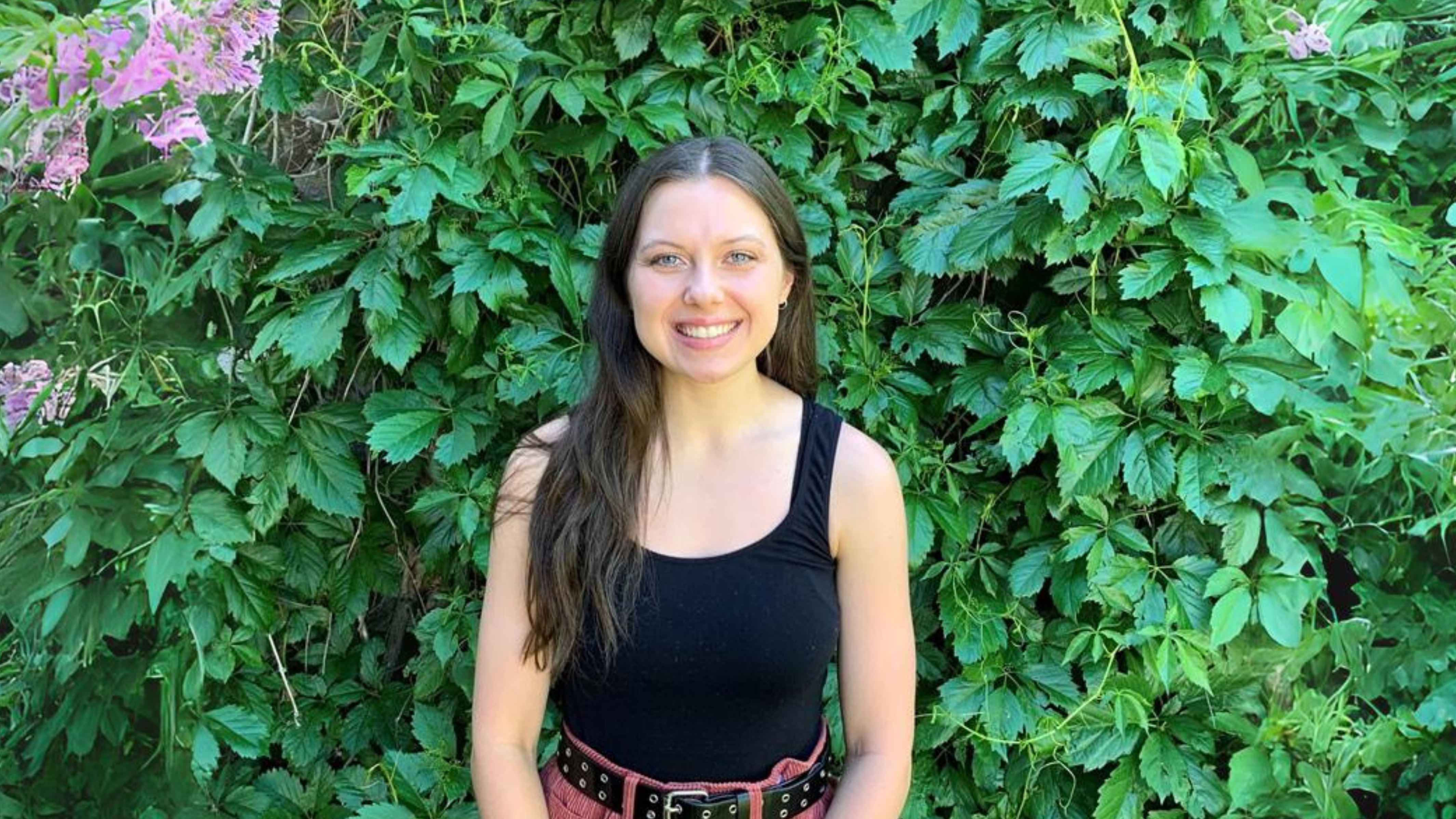 A young woman with long brown hair is smiling while standing in front of a lush green wall of leaves and plants. She is wearing a black sleeveless top and a belt with a red skirt. The vibrant greenery behind her creates a natural and serene backdrop.