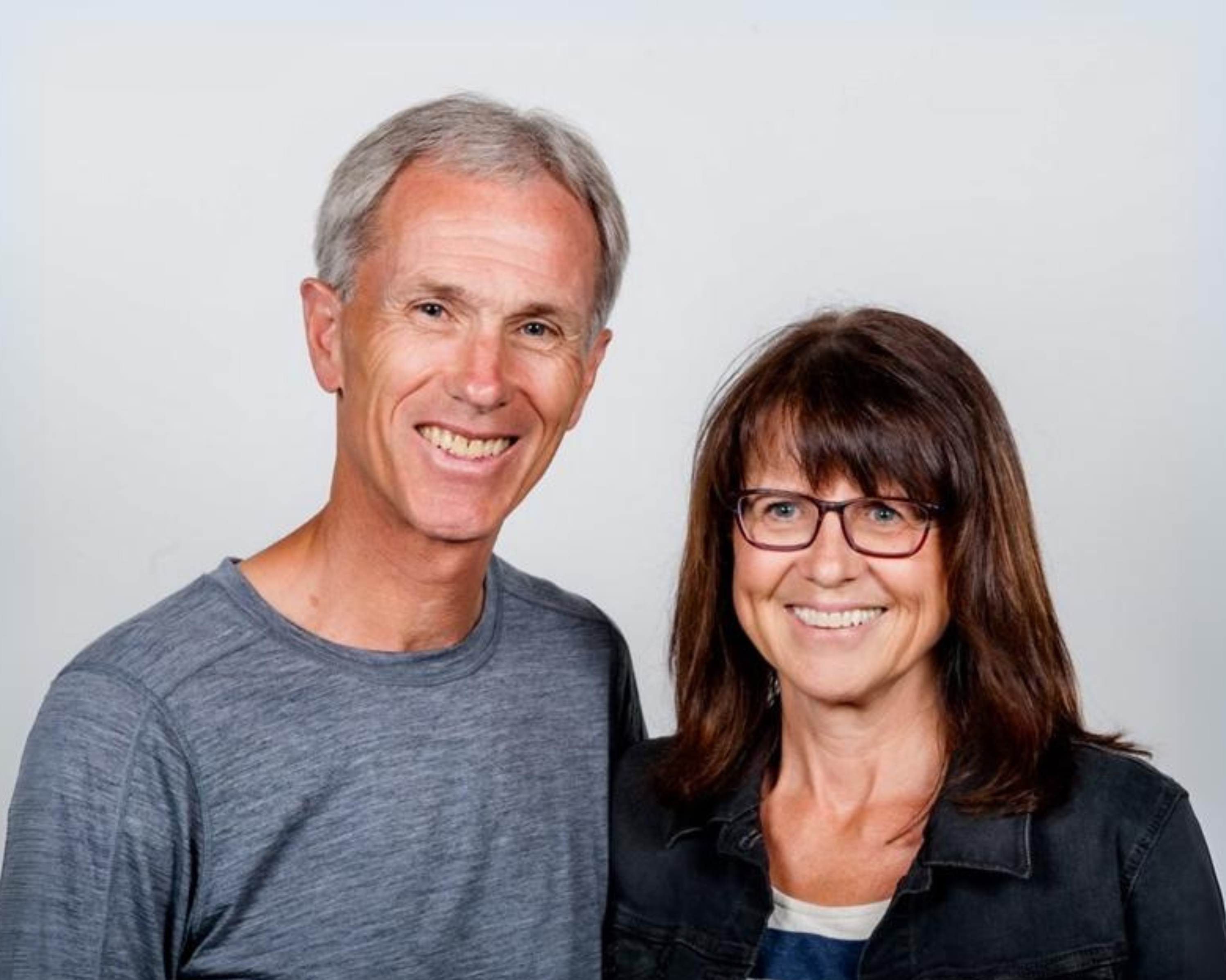 A smiling couple, with the man on the left wearing a grey t-shirt and the woman on the right wearing glasses and a black jacket, standing close together against a plain light background.