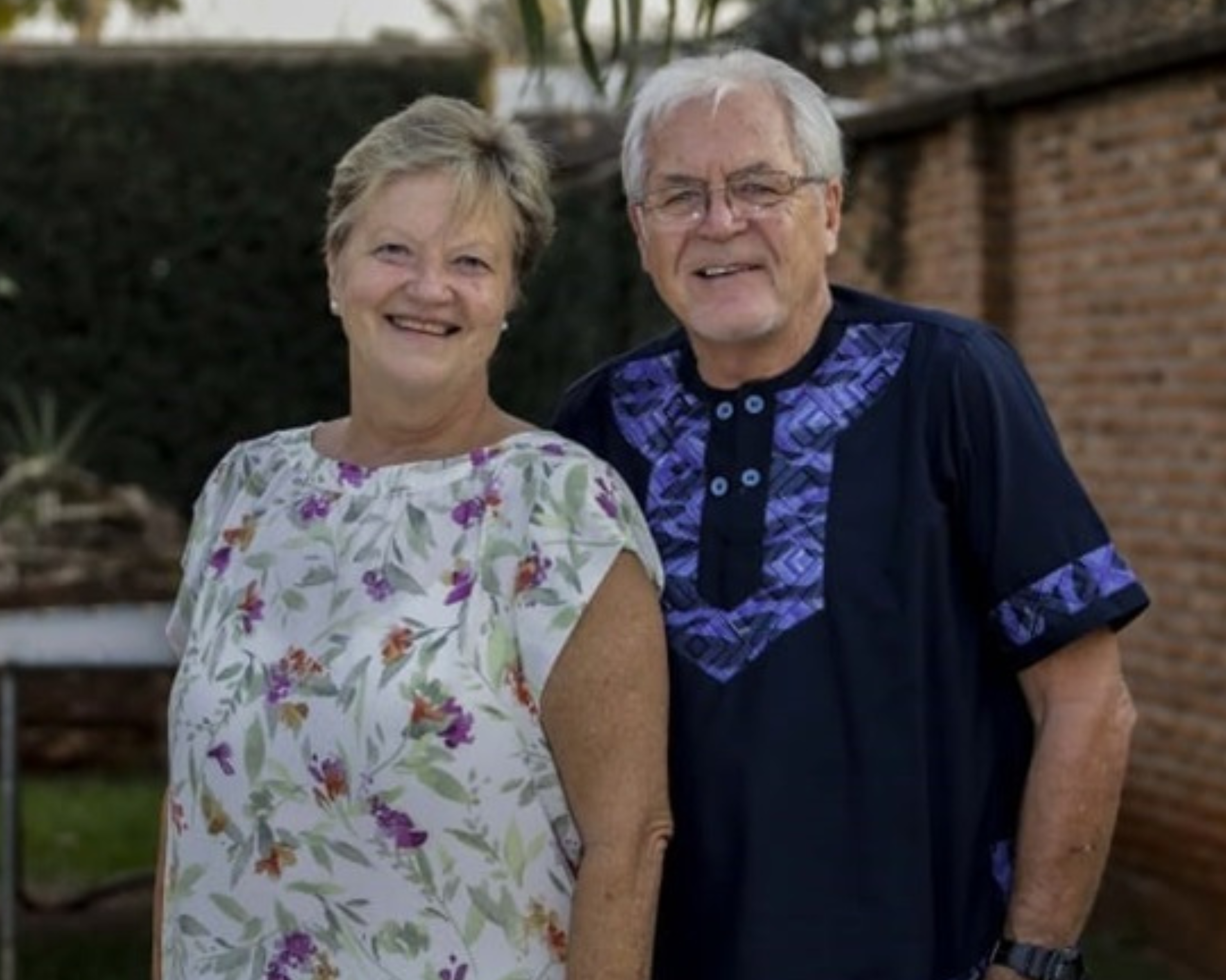 An older couple smiling while standing outdoors in front of a brick wall and some greenery. The woman is wearing a floral patterned blouse, and the man is wearing a dark blue shirt with a purple traditional design. They are both standing close together, looking content and happy.