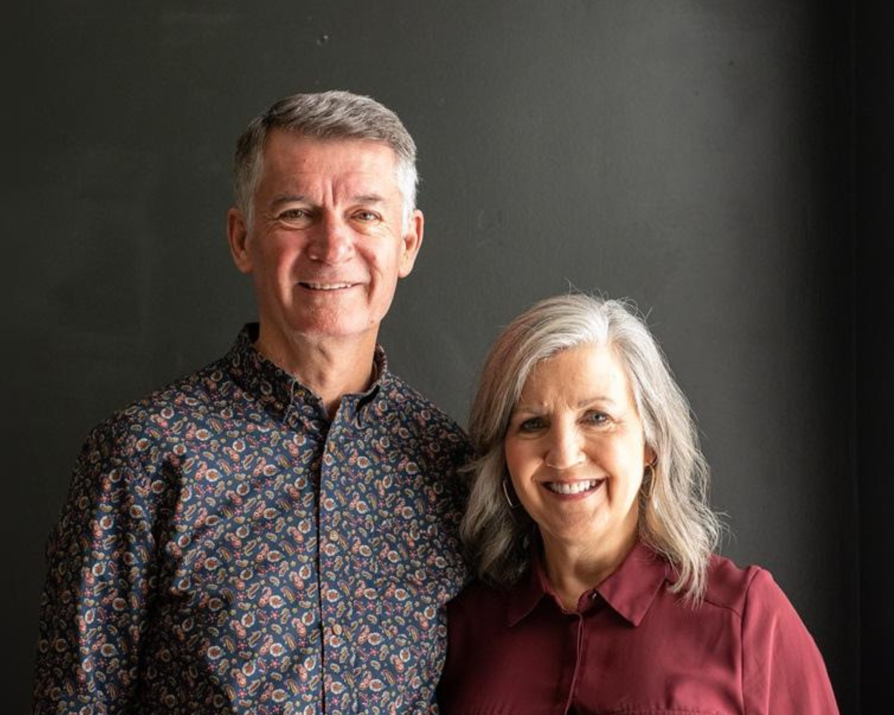 A smiling couple stands side by side against a dark background. The man is wearing a patterned button-down shirt and has grey hair, while the woman, also with grey hair, is wearing a maroon blouse. Both appear happy and relaxed, looking directly at the camera.