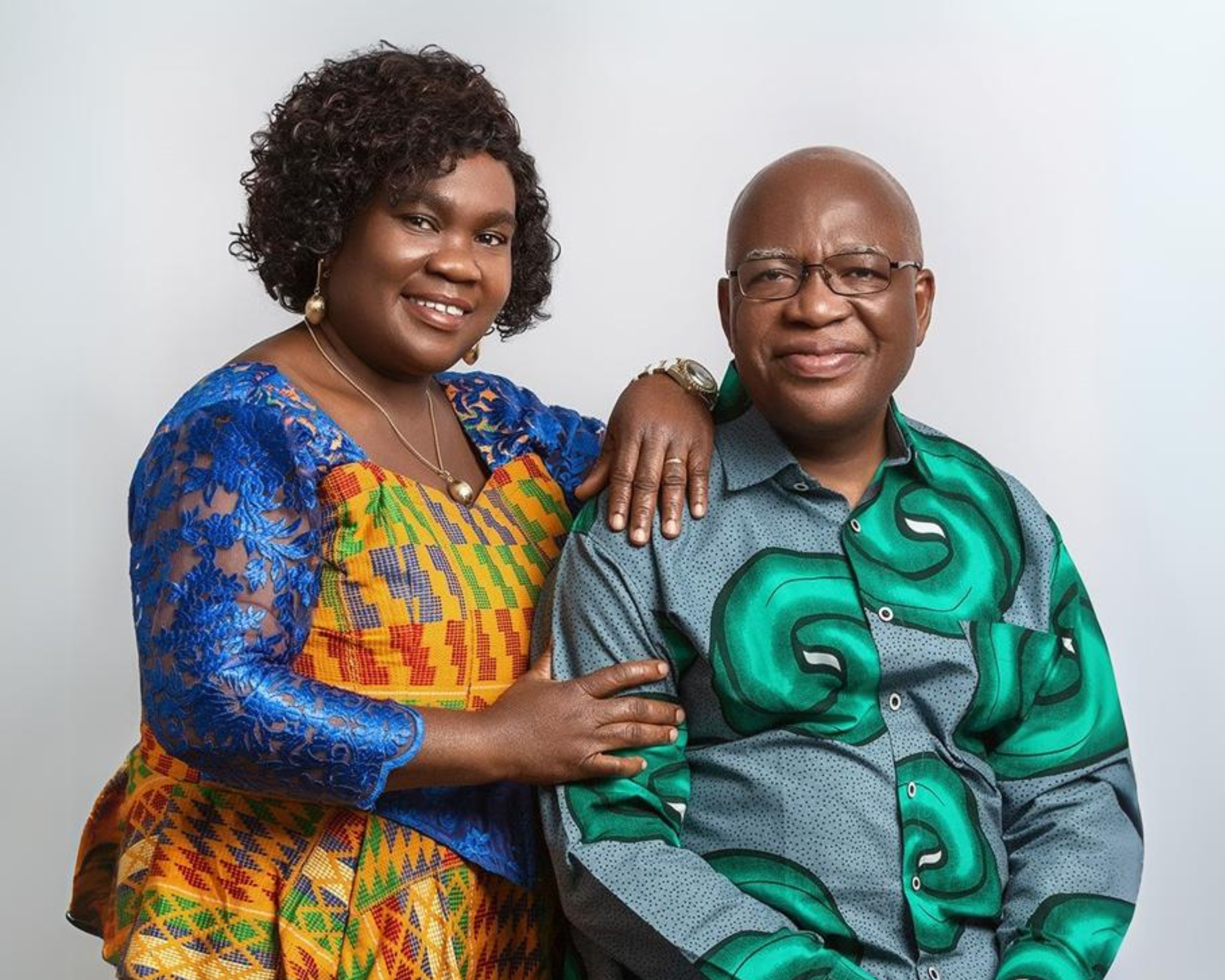 A smiling couple poses for a portrait against a plain white background. The woman, on the left, has curly dark hair and is wearing a colourful, patterned dress with blue lace sleeves. She has her arm affectionately resting on the shoulder of the man beside her. The man, on the right, is wearing glasses and a green patterned shirt. Both of them are smiling warmly at the camera.