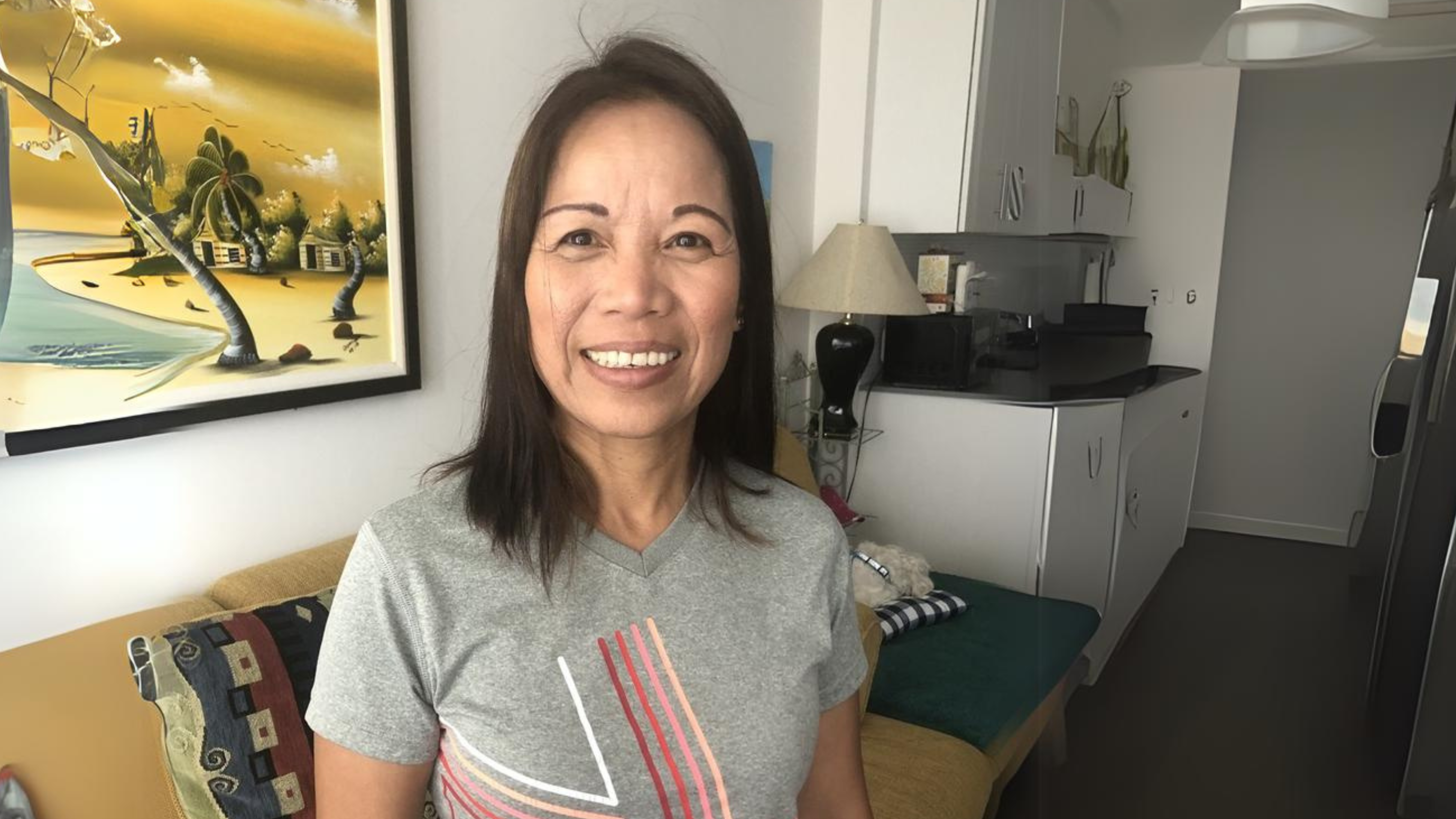 A woman with shoulder-length dark hair is smiling while standing in a cozy, modern living space. She is wearing a gray t-shirt with a colorful design. Behind her, there is a couch with a patterned throw, a lamp, and a kitchen area with white cabinets. A colorful painting of a beach scene hangs on the wall, adding warmth to the room. The overall atmosphere is inviting and comfortable.