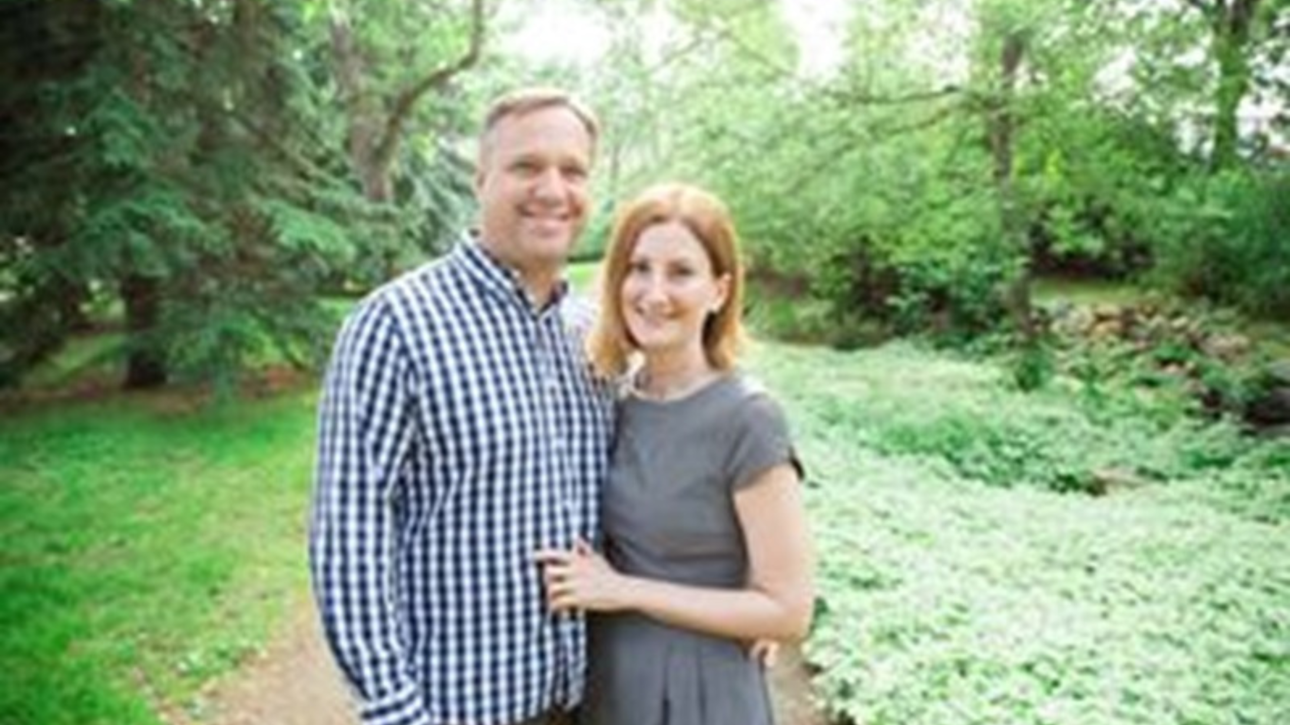 A couple standing closely together on a garden path, surrounded by lush greenery and ground cover. The man is wearing a blue and white checked shirt, and the woman is in a gray dress. They are both smiling, looking directly at the camera, with trees forming a serene backdrop.