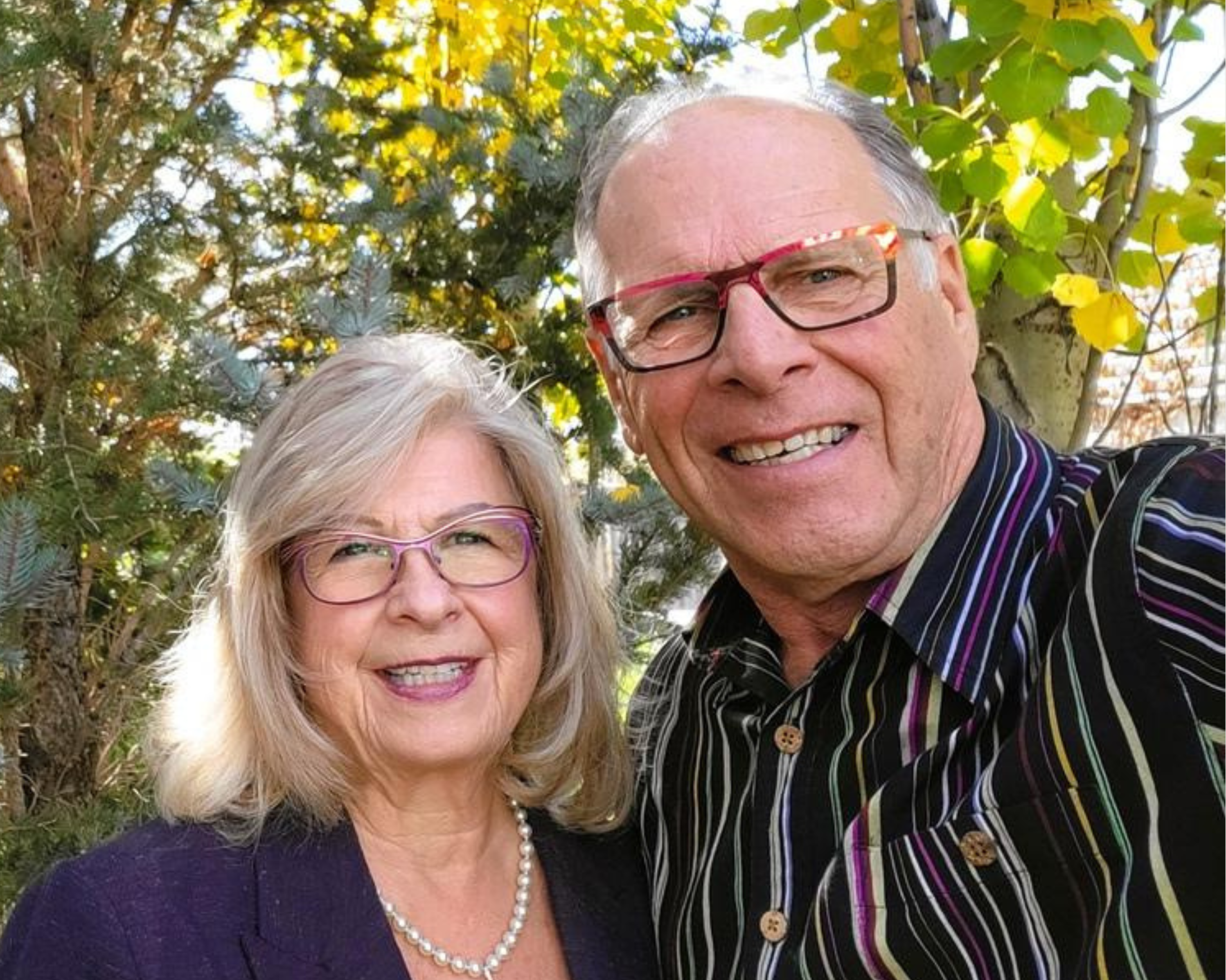 A smiling older couple stands together outdoors in front of a background of trees with autumn leaves. The woman has shoulder-length blonde hair, wears glasses, a pearl necklace, and a dark blazer. The man has short grey hair, wears glasses with red frames, and is dressed in a striped shirt. They both appear happy and relaxed, enjoying the natural surroundings on a sunny day.