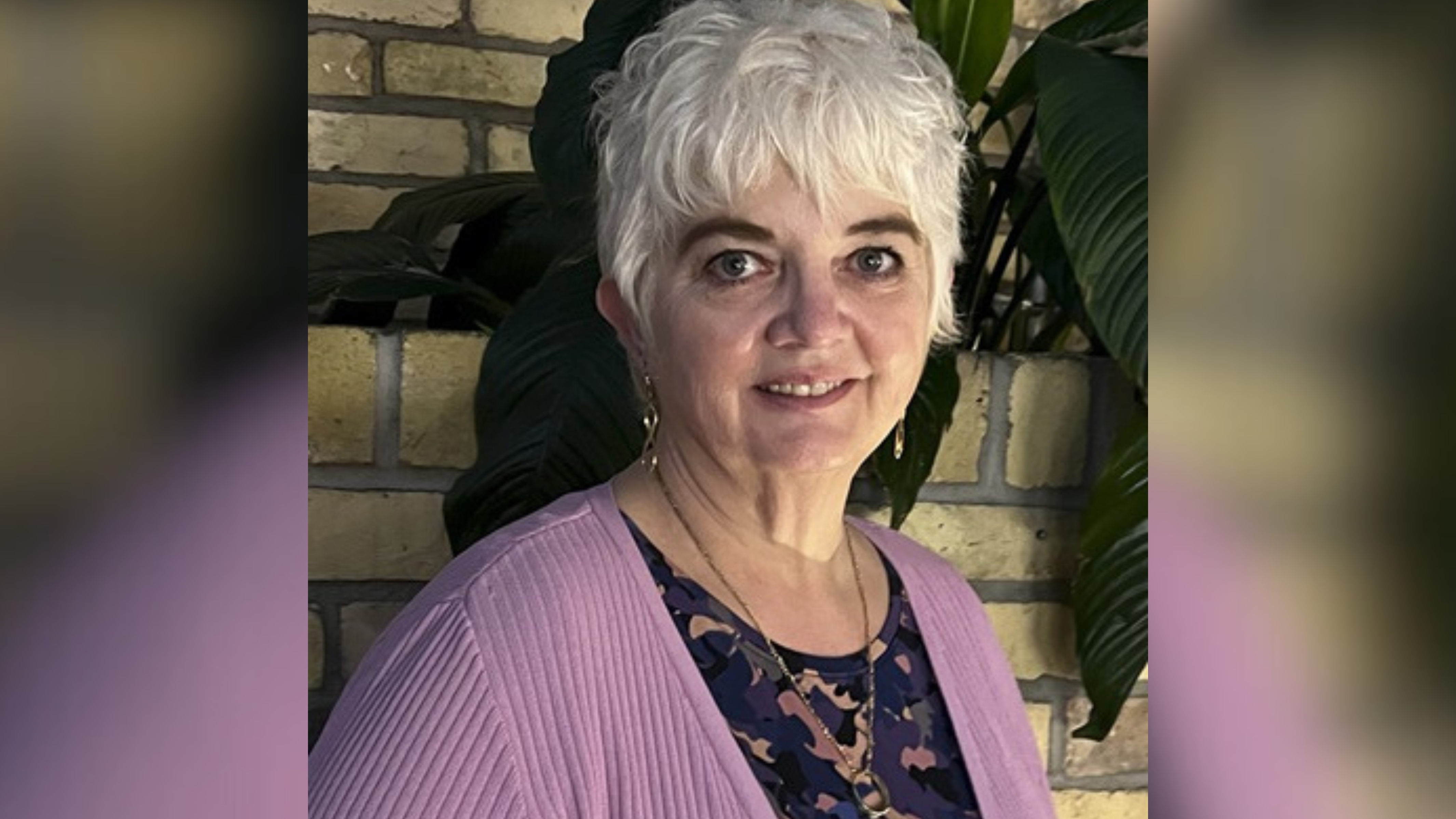 Portrait of an elderly woman with short white hair, smiling gently at the camera. She is wearing a lavender cardigan over a navy and pink floral blouse. The backdrop features a brick wall and lush green plants, adding a warm and inviting feel to the image.