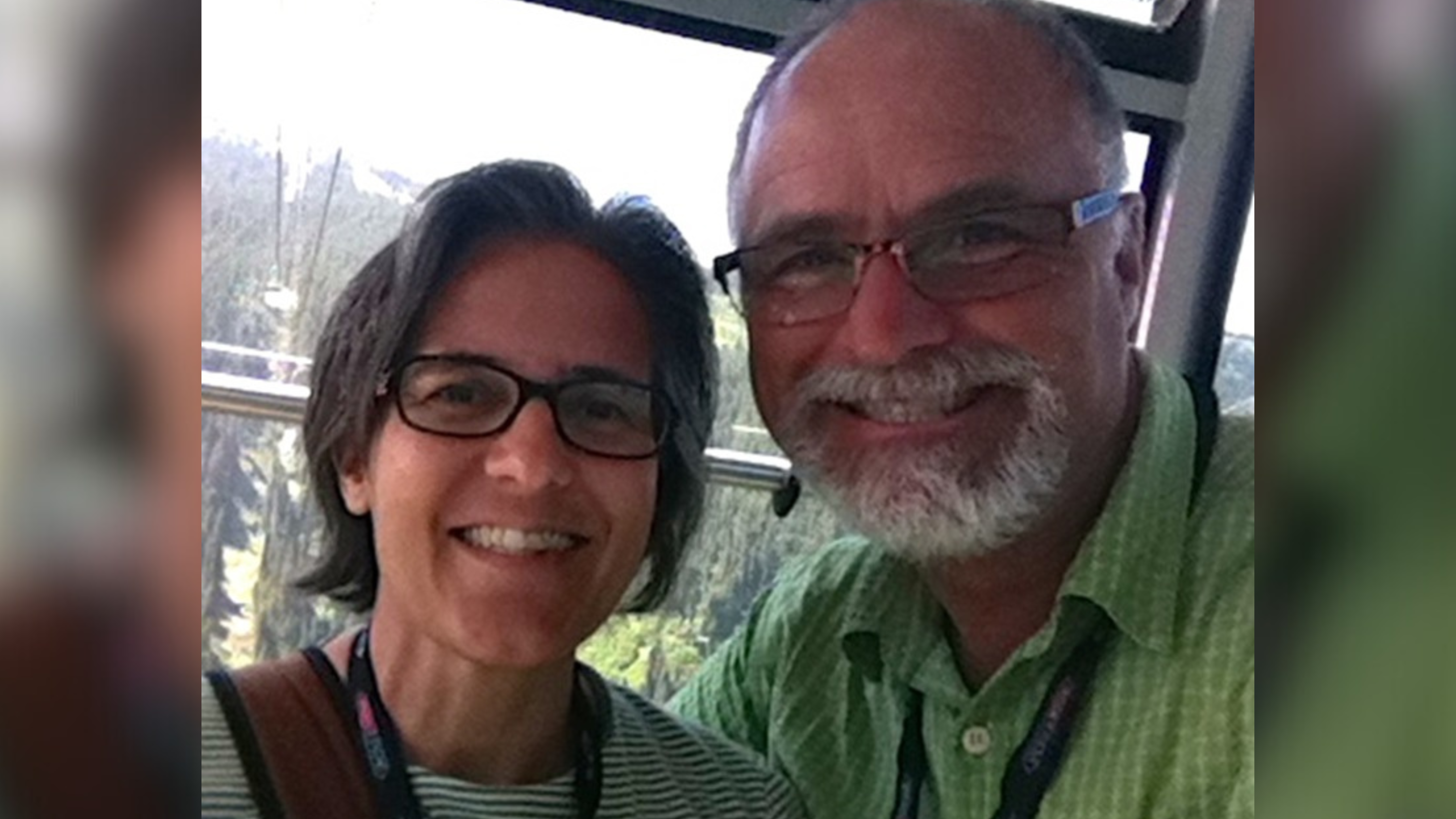 A close-up selfie of a middle-aged couple, a man and a woman, smiling at the camera inside a gondola or cable car. The man is wearing a green striped shirt and glasses, and the woman is wearing a striped shirt and glasses as well. They both wear lanyards, suggesting they might be at a conference or similar event. The background shows a hint of a natural landscape through the window of the cable car.