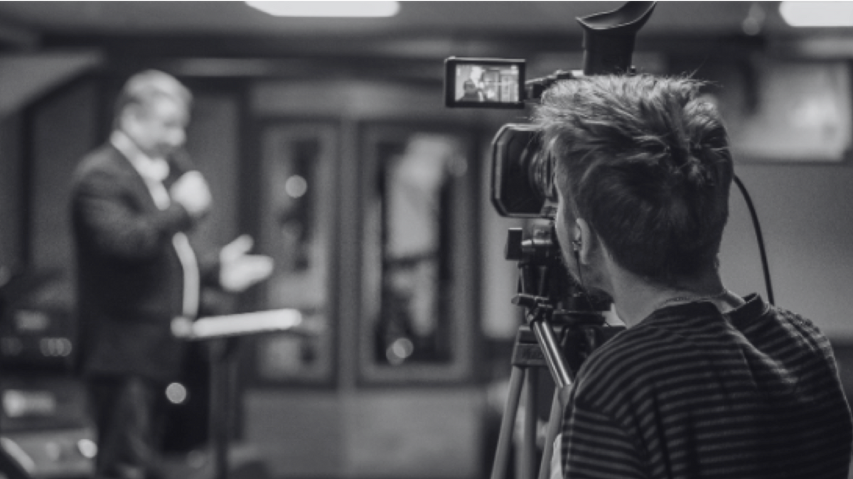 black and white photo of person recording man speaking behind a camera