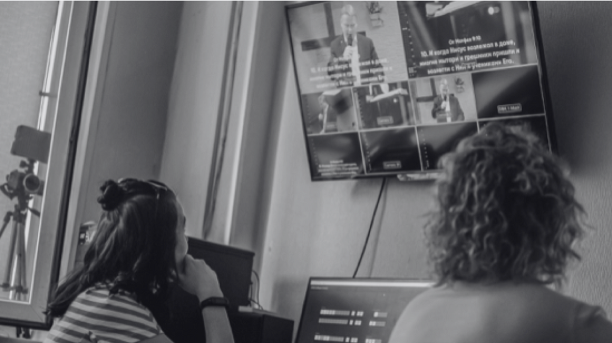 black and white photo of two production individuals looking at a live screen screen of a man speaking on computer monitor and TV screen