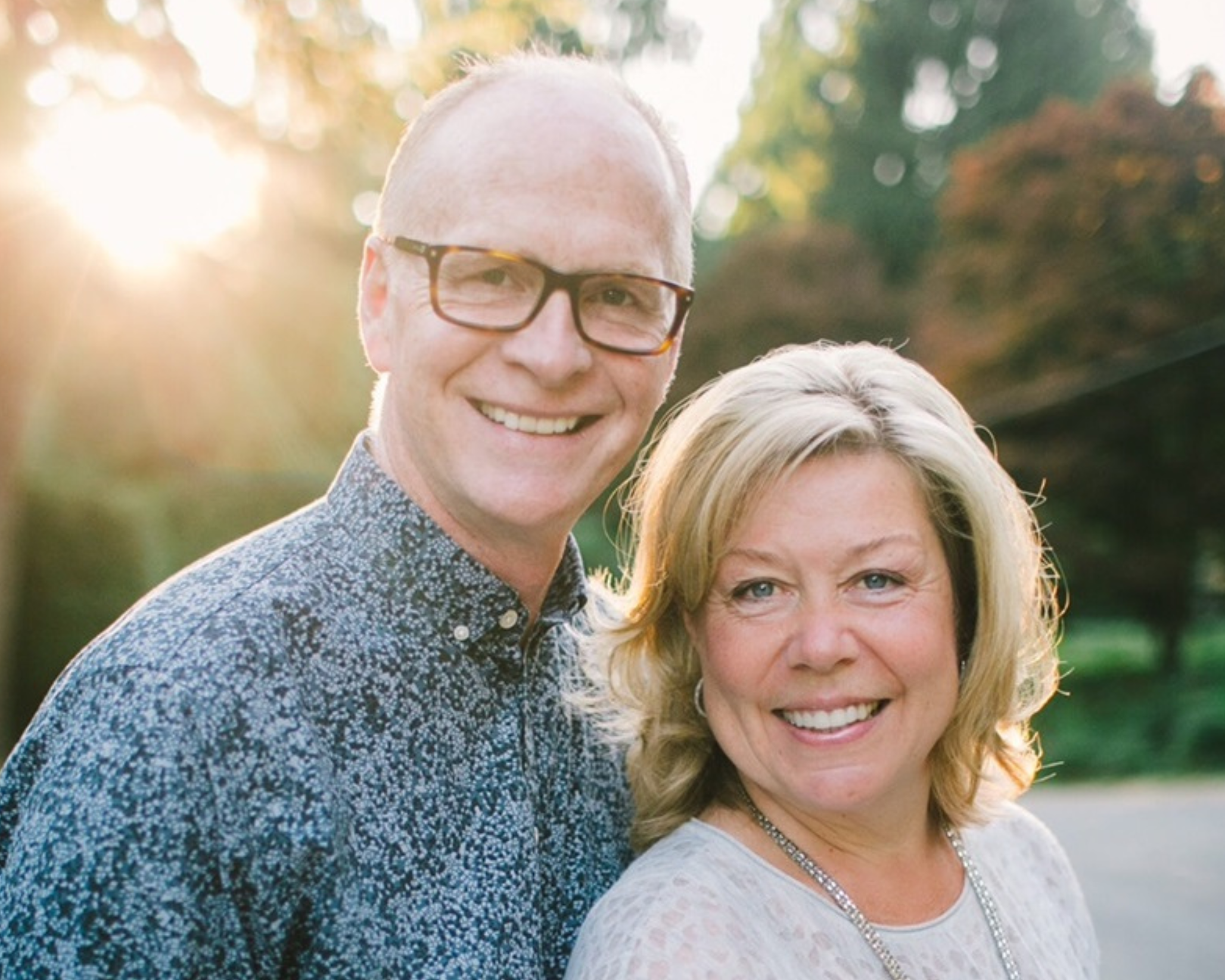Portrait of a middle-aged couple posing outdoors at sunset. The man, wearing glasses and a patterned blue shirt, and the woman, in a light patterned sweater with a long necklace, are smiling warmly. The background is softly blurred with trees and the glow of the sunset enhances their facial features.