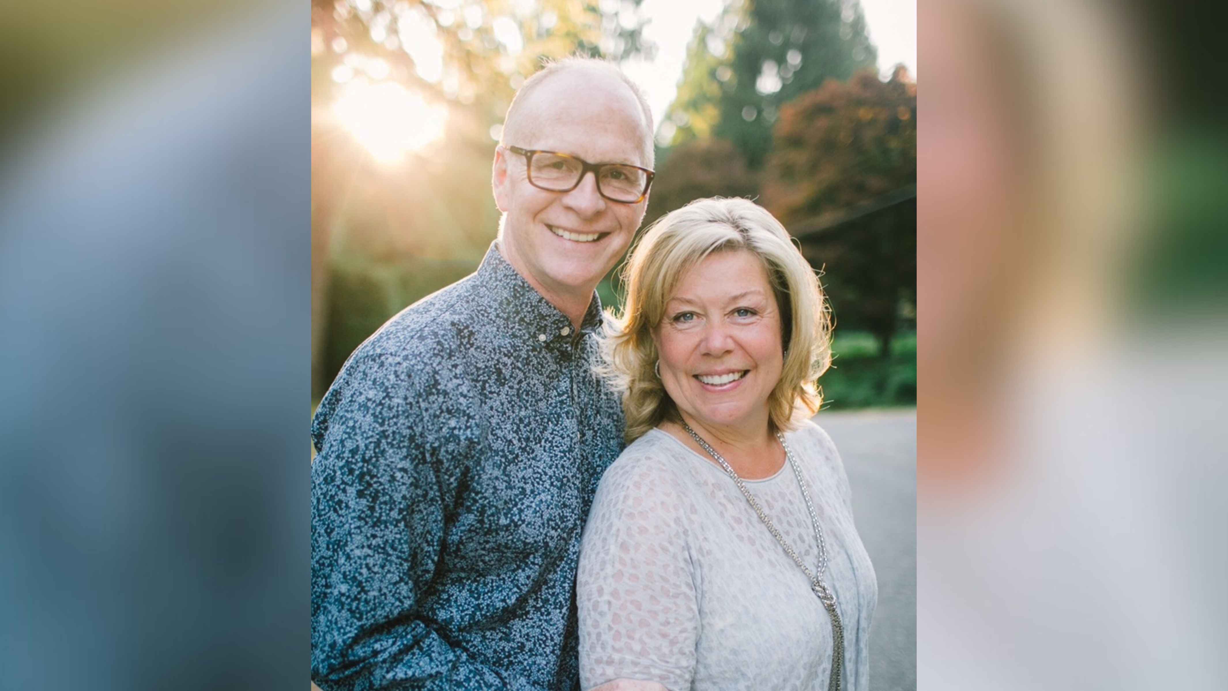 Portrait of a middle-aged couple posing outdoors at sunset. The man, wearing glasses and a patterned blue shirt, and the woman, in a light patterned sweater with a long necklace, are smiling warmly. The background is softly blurred with trees and the glow of the sunset enhances their facial features.