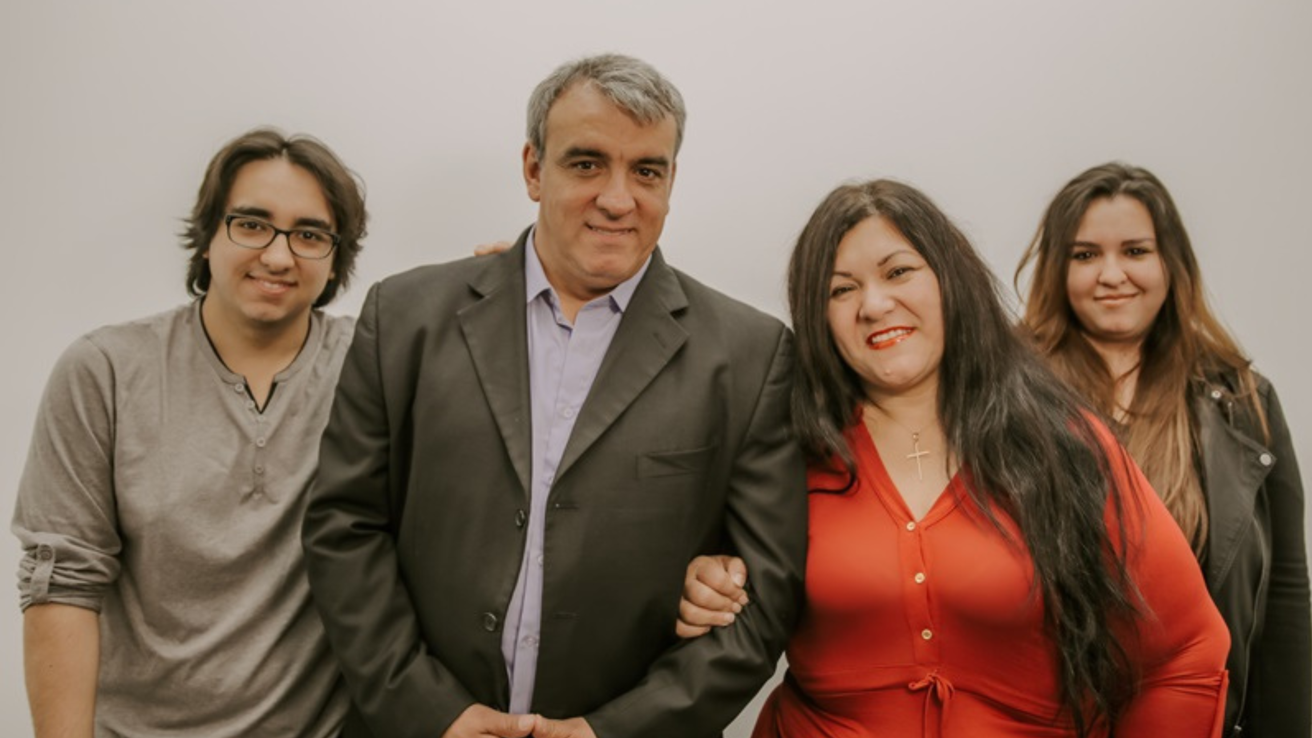 Portrait of a family consisting of four members standing closely together against a neutral background. From left to right: a young man with shoulder-length hair wearing a gray shirt, a middle-aged man in a dark suit and blue shirt, a woman in a bright red blouse, and a young woman in a black jacket. All are smiling warmly at the camera.