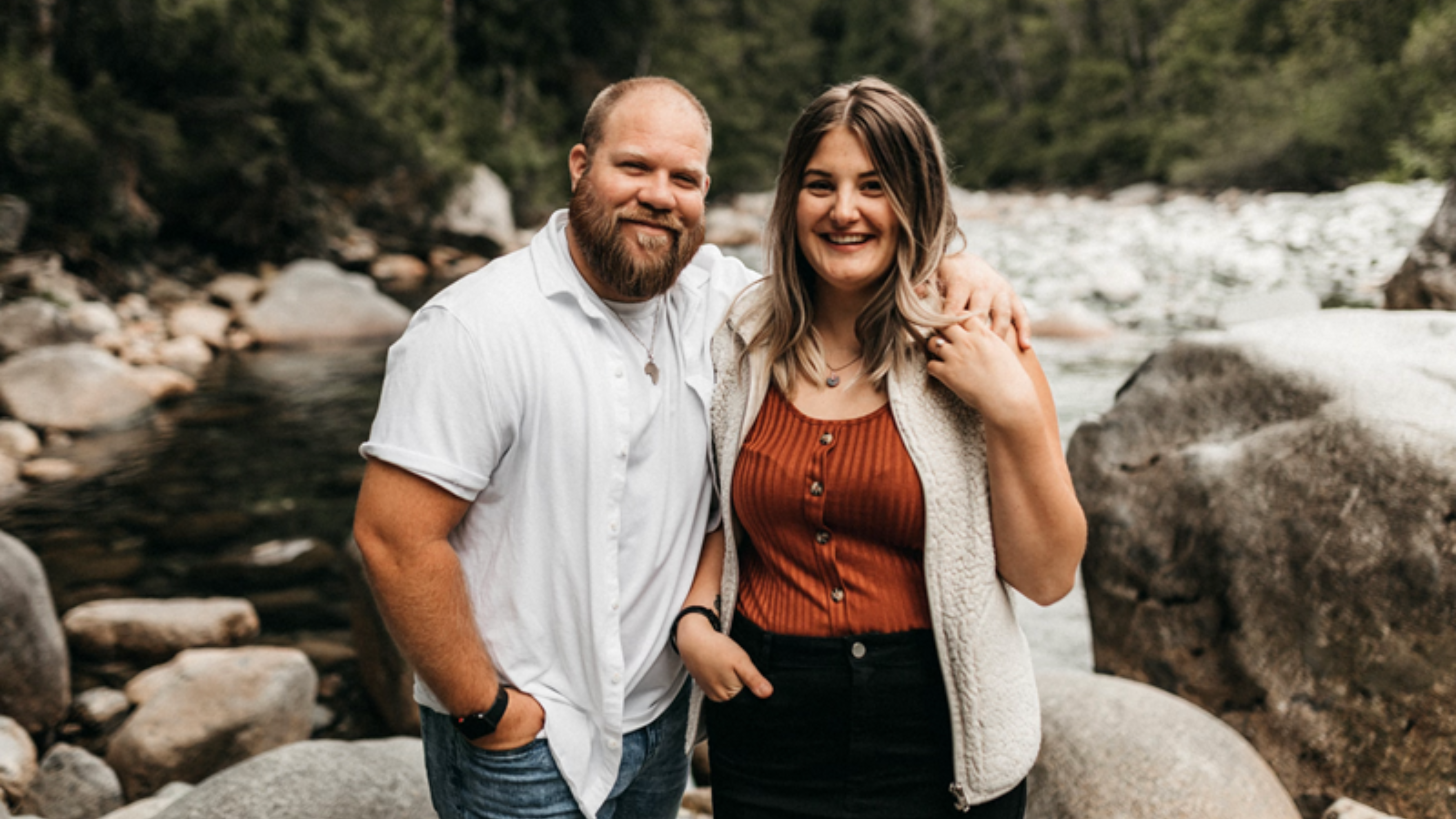 A man and a woman standing together on a rocky riverbank, smiling and posing for the camera. The man is wearing a white button-up shirt and jeans, and the woman is dressed in a rust-coloured top, black skirt, and a light cardigan. They are surrounded by a natural landscape with a flowing river and forested background.