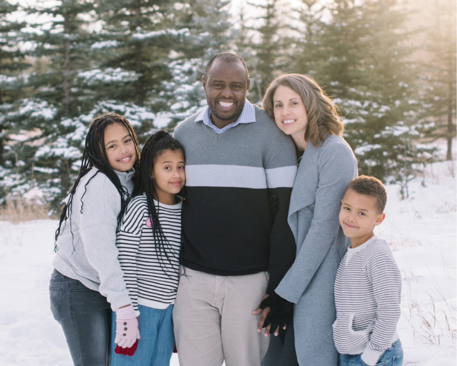 photo of couple and three young children smiling at camera behind scenery of snow and trees.