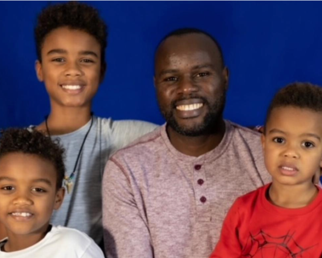 photo of man and three young boys smiling at camera behind blue wall.