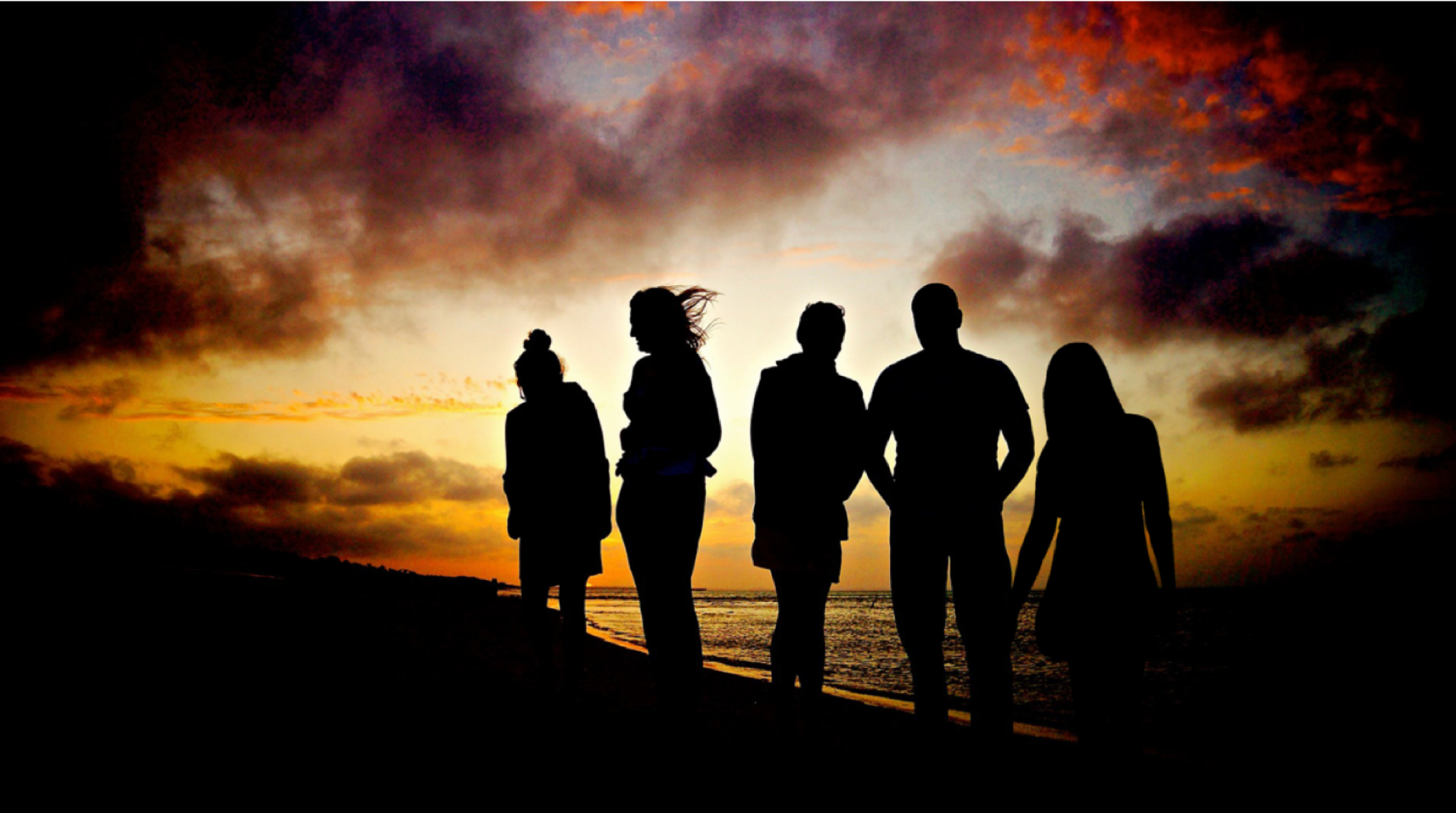 Silhouettes of five people standing together on a beach at sunset, with dramatic clouds and vibrant colours in the sky, creating a serene and reflective atmosphere.