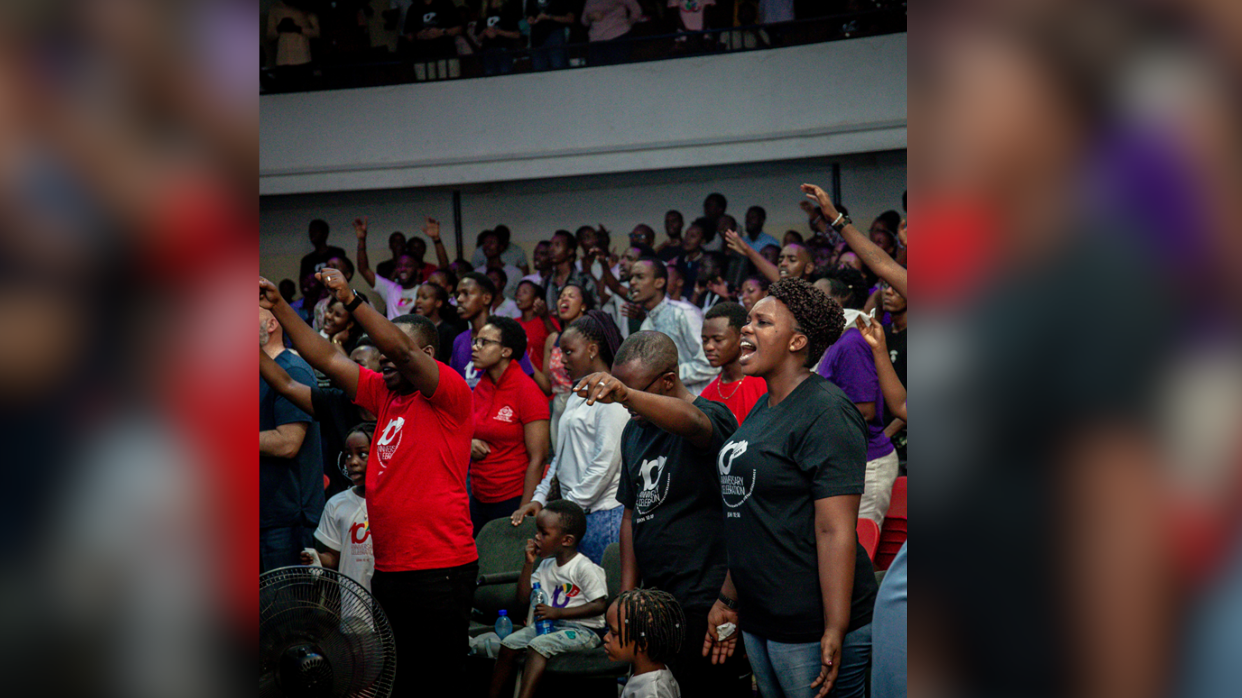 A diverse group of people engaged in a worship service, with some raising their hands and singing passionately. The crowd includes both adults and children, all gathered in a large indoor space.