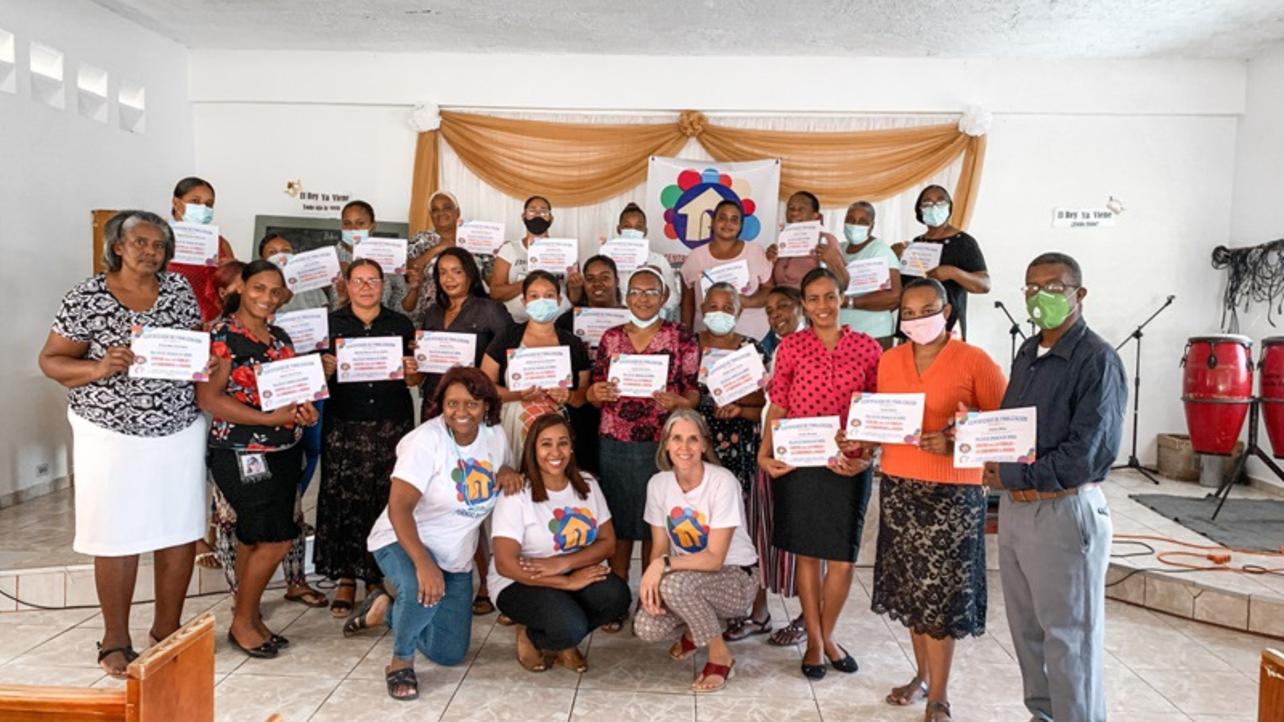 A large group of people standing inside a church, holding certificates. Some are wearing white T-shirts with a colourful logo on them, and others are dressed in various outfits. The church's wall behind them has the words "El Rey Ya Viene" written on it, and some people in the group are wearing face masks.