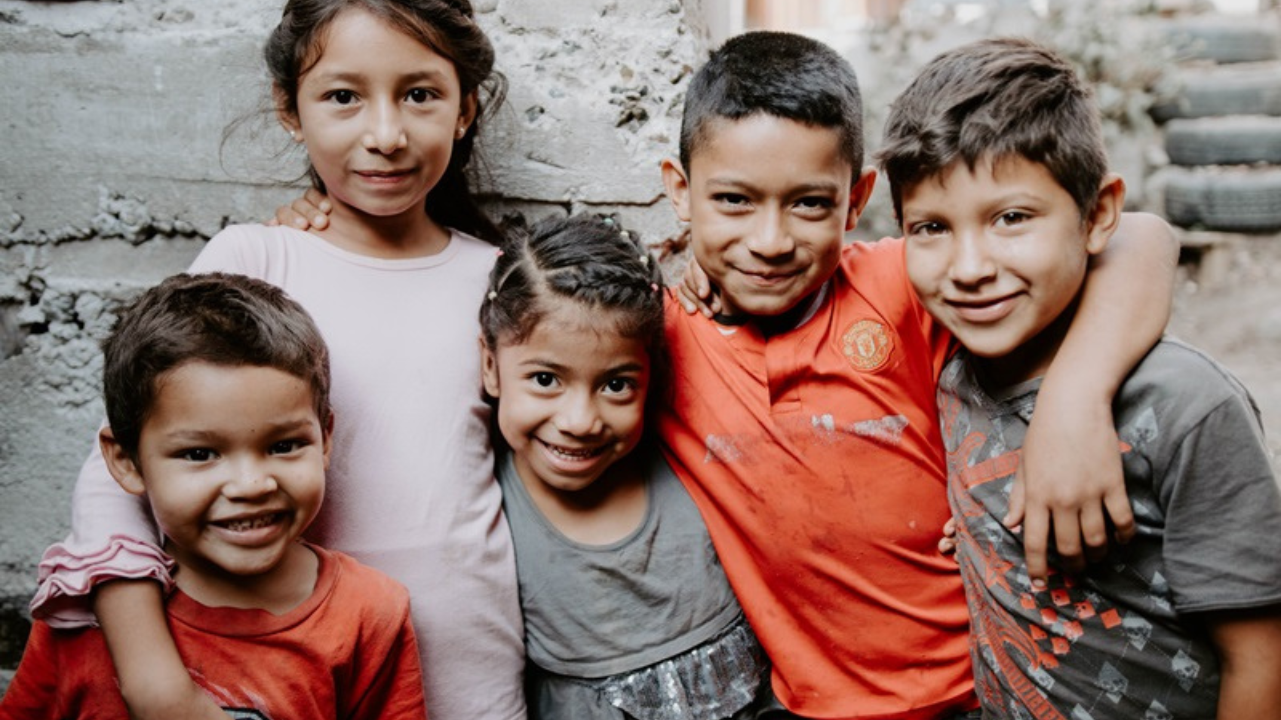 Five smiling children stand close together, with their arms around each other. They are dressed casually and are standing against a rough, textured wall in an outdoor setting.